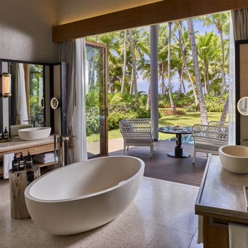 Luxuriöses Badezimmer mit freistehender weißer Badewanne, die auf eine offene Terrassentür blickt, die zu einem Außenbereich mit Sitzgelegenheiten umgeben von Palmen und grüner Vegetation führt.