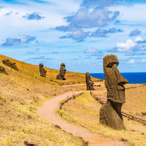 Multiple Moai statues along a dirt path on Easter Island with the ocean and cloudy sky in the background.