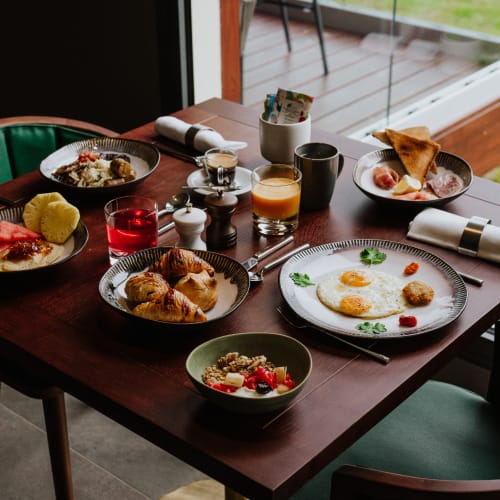 Ein Holztisch mit Frühstück, darunter Croissants, Spiegeleier, Müsli mit Früchten, Wassermelonenstücke, Toast und Getränke wie Wasser und Orangensaft, nahe einem Fenster mit grünen Stühlen.