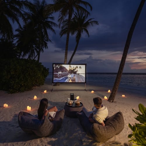 Ein Paar sieht sich bei Sonnenuntergang auf einer tropischen Insel am Strand einen Film bei einem Open-Air-Kino an, umgeben von Palmen und Kerzen.
