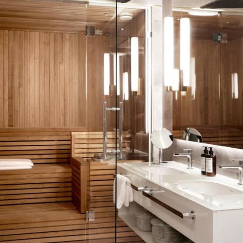 A modern bathroom with a double sink vanity, mirror with vertical light strips, and a glass door leading to a wooden sauna room with a towel on the bench.