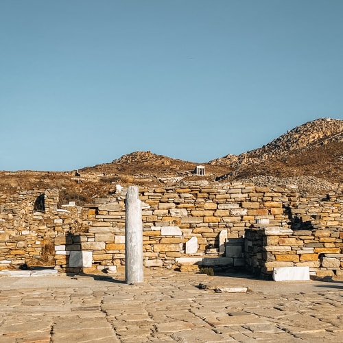 Ancient stone ruins on Delos Island near Mykonos, featuring a single standing marble column against a backdrop of rocky hills under a clear blue sky.