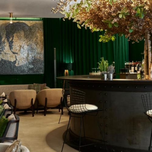 Eine stilvolle Hotelbar mit grüner Dekoration, Holzbar und Sitzbereich mit Ohrensesseln sowie Drahtstühlen.