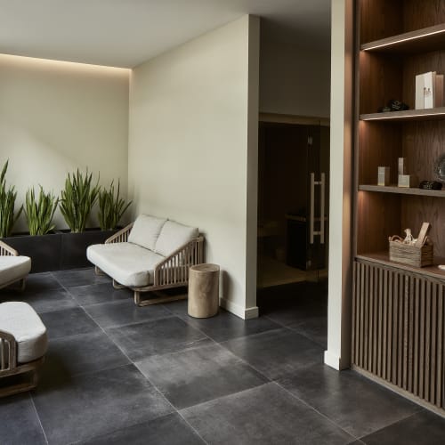 Moderner Spa-Wartebereich mit drei gepolsterten Holzstühlen, einem kleinen Holzbeistelltisch, einem Pflanzkübel mit hohen grünen Pflanzen in der Ecke und einem beleuchteten Holzregal.