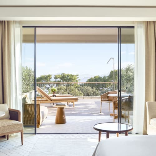 Modern resort room with a bed, sofa, armchairs, and a round table, overlooking a balcony with sun loungers and lush greenery beyond.