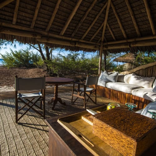 Ein schattiger Strandloungebereich mit Holz-Möbeln und einer gepolsterten Sitzgelegenheit unter einem Strohdach, mit Blick auf den Strand und den Ozean.