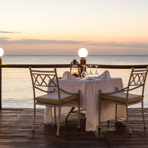 Ein romantisches privates Abendessen auf einem Holzdeck mit Blick auf den Ozean bei Sonnenuntergang, mit einem runden Tisch in weißem Tuch, umgeben von vier Stühlen, und Kerzen im Licht.