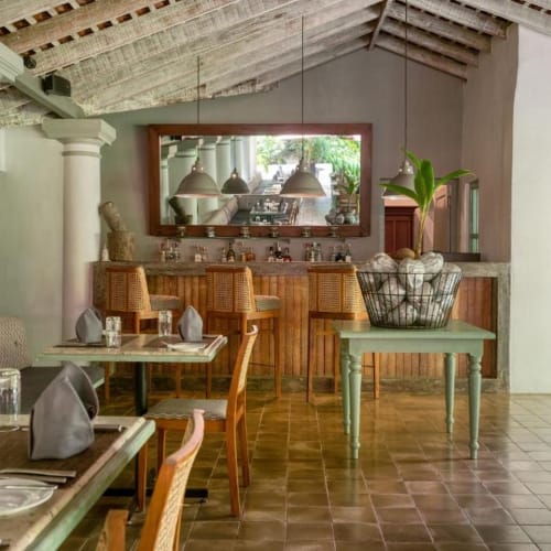 Ein rustikales Restaurant mit gemustertem Holzdecken, weißen Säulen, einem großen Wandspiegel, einem kleinen Tisch mit Pflanzen und mehreren Tischen mit Besteck und Gläsern.