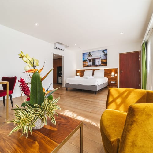 Ein modernes Hotelzimmer mit einem ordentlich gemachten Bett, einem Holztisch mit einer lebhaften Blumenanordnung, einem gelben Sessel und großen Fenstern mit grünen Vorhängen, durch die Sonnenlicht fällt.