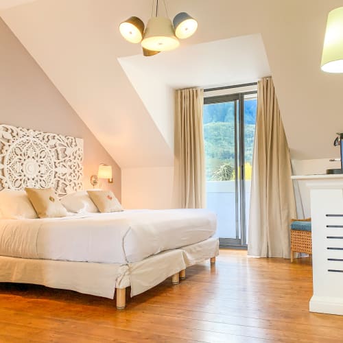 Helles Deluxe-Schlafzimmer mit großem Bett, kunstvoll verziertem Kopfteil, Holzboden und Glastür zum Balkon mit Bergblick.