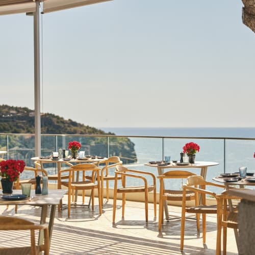 Außenrestaurant-Terrasse mit Holzstühlen und Tischen, geschmückt mit roten Blumen, mit Blick auf das Meer und eine Küstenklippe unter klarem Himmel.