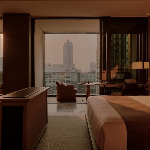 Ein luxuriöser Hotelzimmer mit einem großen Bett, Wandpaneelen aus Holz und einem Sitzbereich mit Blick auf die Stadt durch große Fenster.