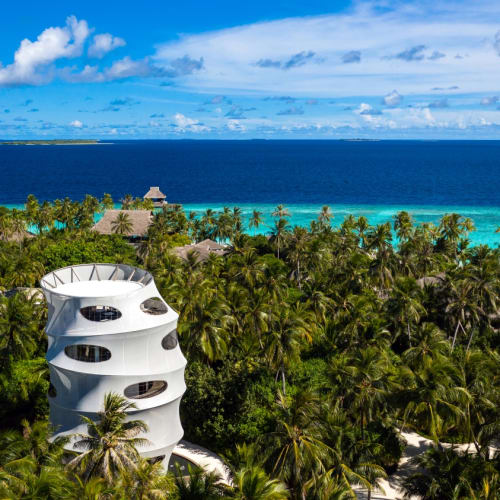 Hoher moderner weißer Spiralturm umgeben von dichtem Palmwald mit türkisfarbenem Ozean und blauem Himmel im Hintergrund.