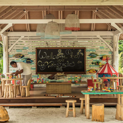 Offener Kinderclub-Bereich im Four Seasons Resort Seychelles auf der Insel Desroches mit Holztischen, Hockern, Spielzeug und einer Tafel mit der Aufschrift 'Welcome to Desroches'. Ein Mitarbeiter ordnet Gegenstände auf einem Tisch unter einem Holzpavillon inmitten von Grünflächen.