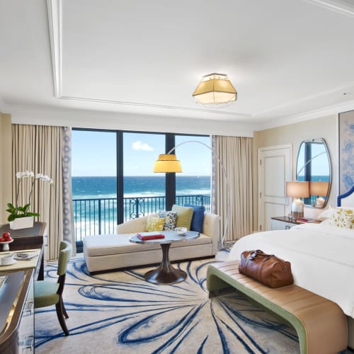 Luxurious hotel room with a large bed, a sitting area near the window overlooking the ocean, and elegant decor.