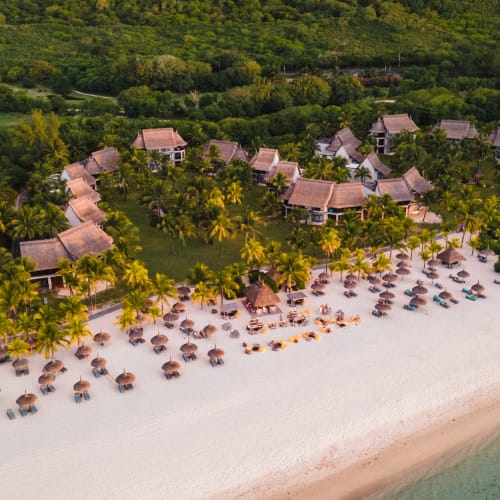 Aerial view of a tropical beach resort with villas surrounded by lush greenery and rows of umbrellas and sun loungers on a sandy beach beside clear ocean water.
