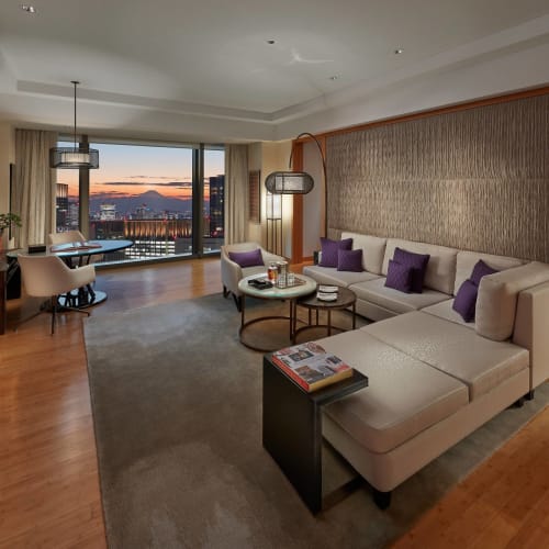 Spacious living room in a high-rise apartment with large windows showing a cityscape at sunset, featuring a white sectional sofa, purple cushions, a coffee table, and a work desk.