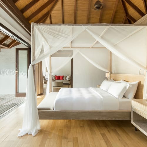 Eine helle Strandvilla mit einem vierpfostigen Bett, das mit weißem Stoff drapiert ist, Holzwände und eine Schiebetür, die auf eine offene Terrasse mit Liegestühlen führt.