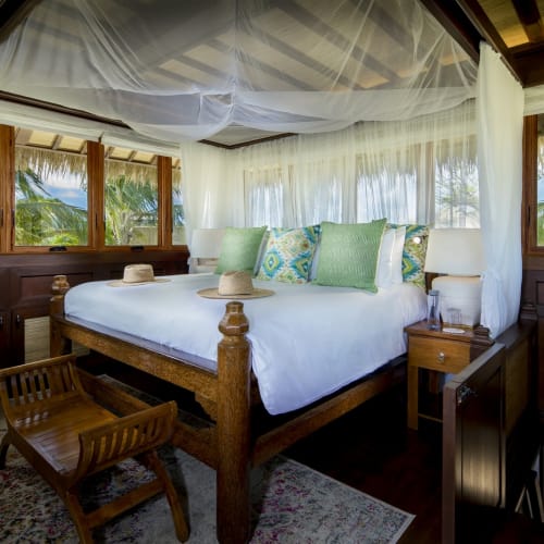 Luxurious bedroom with large bed, colorful pillows, and mosquito net, surrounded by large windows showing lush outdoor greenery.