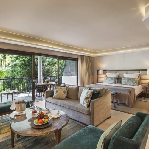 Modern hotel suite with a large bed, a sitting area with a sofa and armchairs, a coffee table with fruit and drinks, and a balcony overlooking lush greenery.