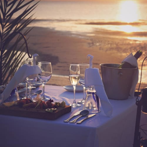 Elegant beachside dinner setup with wine glasses, a bucket of champagne, and a wooden platter of food against the backdrop of a sunset on the beach.