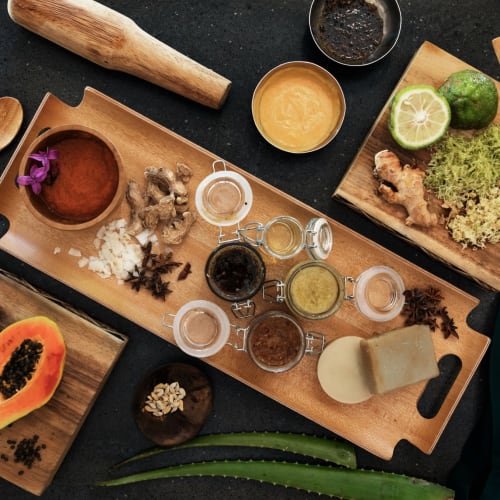 Verschiedene Zutaten und Küchenutensilien sind auf Holztafeln und einer schwarzen Oberfläche arrangiert, einschließlich Früchte, Gewürze, Öle und Holzutensilien.