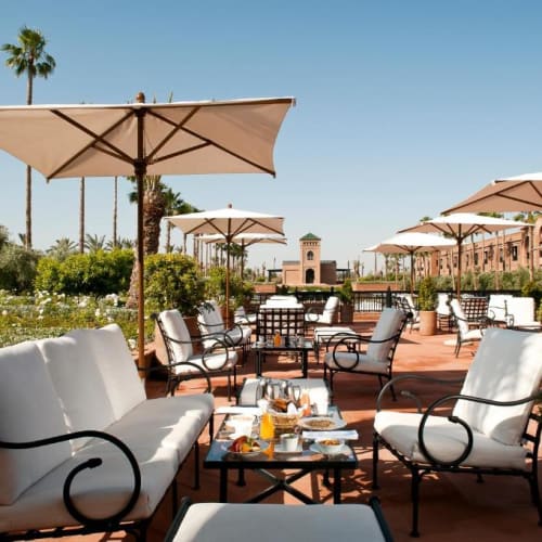 Außenbereich mit weißen gepolsterten Stühlen und Tischen unter großen beigefarbenen Sonnenschirmen bei einem Hotel in Marrakech, Marokko.