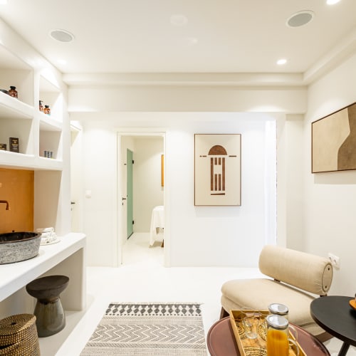 Bright and modern spa interior with white walls, two stone washbasins on a countertop with brass faucets, a seating area with a round black table holding a fruit basket and drinks, and minimalist framed artwork on the walls.