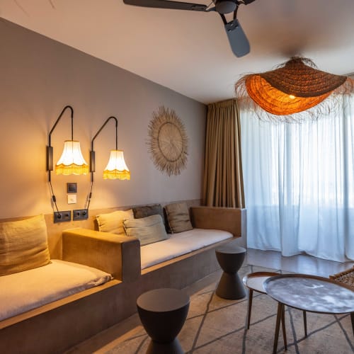 Gemütliches Wohnzimmer mit eingebauten beigen Sofas, weichen Kissen und zwei hängenden Lampen an der linken Wand, zusammen mit gewebten Wanddekorationen und einer einzigartigen gewebten Deckenlampe neben großen, mit transparenten Vorhängen bedeckten Fenstern.