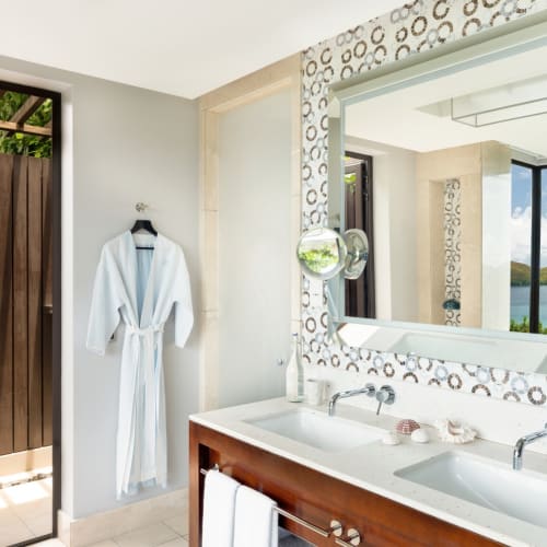 Ein helles Badezimmer mit zwei Waschbecken, großem Spiegel, weißem Bademantel an der Wand und einer offenen Tür, die zu einem Außendusche-Bereich mit Holzzaun und Grünflächen führt.