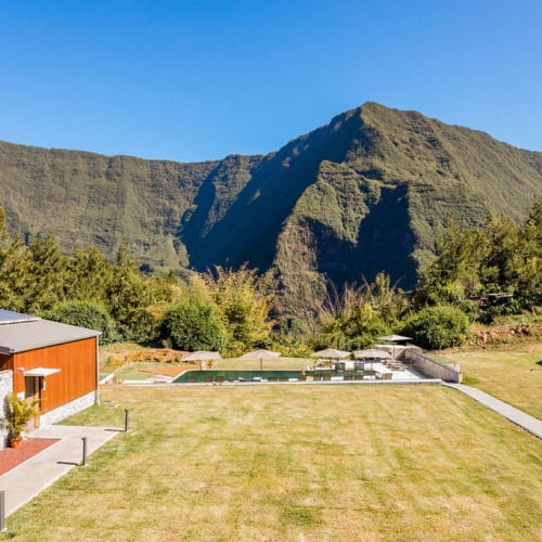 A modern outdoor area with a small pool, surrounded by green lawns and trees, set against the backdrop of large green mountains under a clear blue sky.
