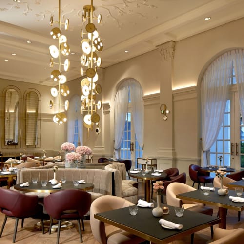 Eleganter Speiseraum mit modernen Kronleuchtern, großen bogenförmigen Fenstern mit Vorhängen und einer Mischung aus Stühlen an dunklen Tischen im Raffles Singapore La Dame de Pic Restaurant.