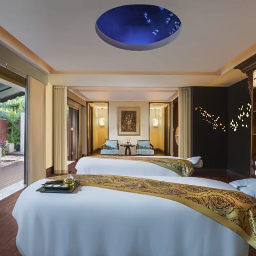 Ein luxuriöser Spa-Behandlungsraum mit zwei Massageliegen, eleganter Einrichtung und Zugang zu einem Außenbereich mit Grünflächen.