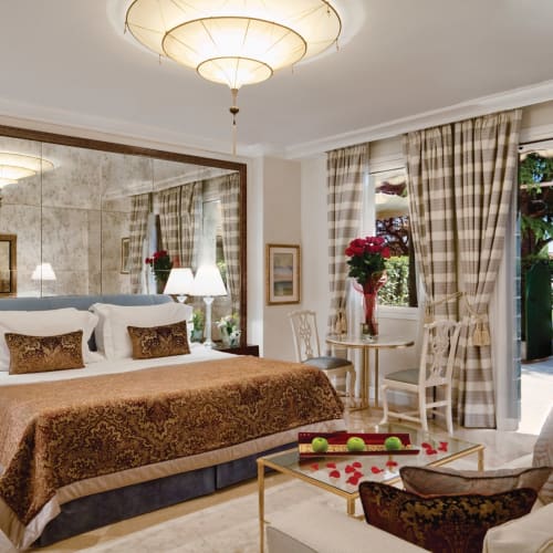 Luxus호텔zimmer mit großem Bett, dekorativen Kissen, einem spiegelflächigen Kopfteil und einer Sitzecke neben Fenstern mit karierten Vorhängen, mit Blick auf den Pool.