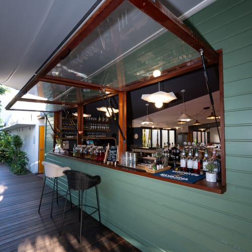 Außenbar mit mintgrünen Holzpaneelen und zwei Hochtischen unter einem geöffneten transparenten Dachfenster, mit Regalen voller Flaschen und Gläser innen und hängenden Pendelleuchten.