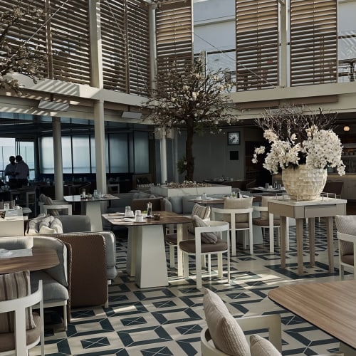 Modernes Restaurantinnere mit gemustertem Fliesenboden, Holztischen und Stühlen mit Kissen sowie einem großen Blumenarrangement unter einer Lamellendecke, die natürliches Licht hereinlässt.