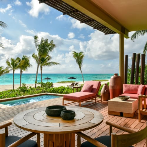 A luxurious beachfront terrace with pink lounge chairs, a wooden dining table, and lush tropical greenery overlooking the beach and ocean.