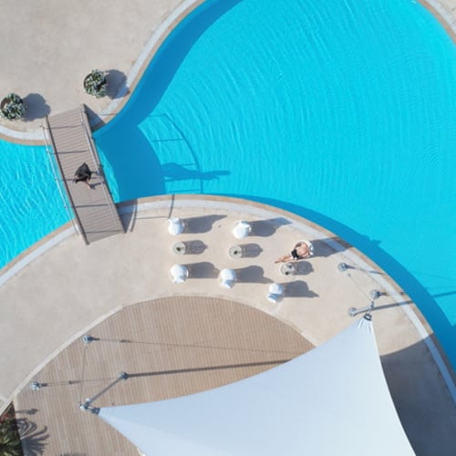 Luftaufnahme eines Hotelpools mit zwei Schwimmbecken, Sonnenliegen, Sonnenschirmen und einem schattigen Zelt in einer modernen Außenanlage.