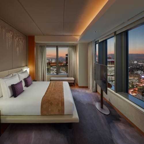 Luxushotelzimmer in Tokio mit großem Bett, Flachbildfernseher und Blick auf die Skyline der Stadt bei Sonnenuntergang.