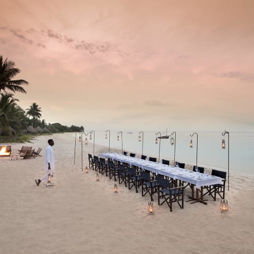 A beautifully set outdoor dining table on a sandy beach at sunset, with palm trees and calm sea in the background.