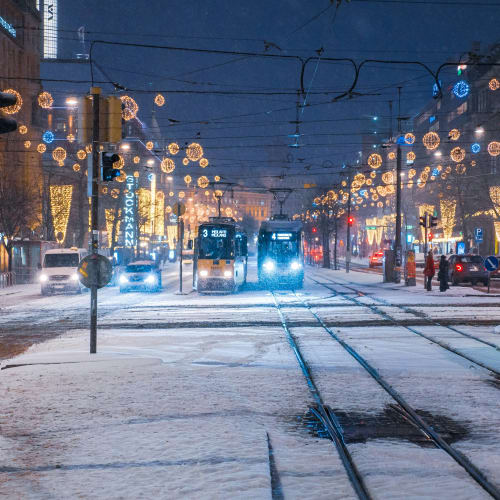 Snow-covered tram tracks in a city street at night with trams approaching, decorated with warm festive lights and illuminated buildings.