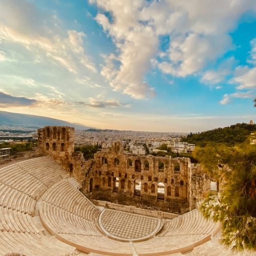 Ancient stone amphitheater with curved seating overlooking the city of Athens under a partly cloudy sky at sunset.