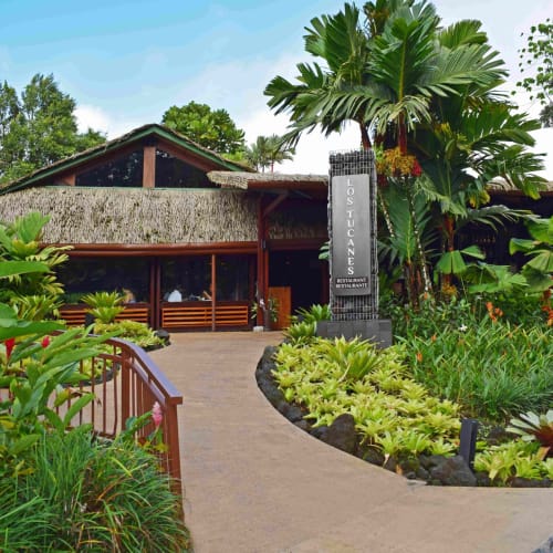 The entrance pathway of Tabacón Thermal Resort and Spa with lush greenery and a building in the background.
