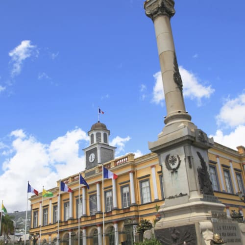 Das historische Rathaus von Saint-Denis, Réunion, mit einem Uhrturm und mehreren Flaggen, darunter die französische Flagge, neben einem hohen steinernen Denkmal mit einer Engelsstatue unter einem strahlend blauen Himmel.