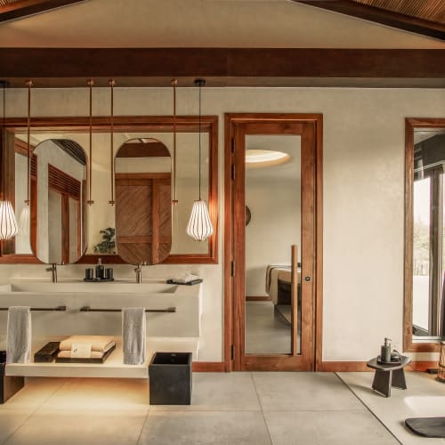 Modernes Badezimmer in einer Honeymoon-Villa mit Doppelwaschbecken, großen Spiegeln, Holzakzenten, hängenden Pendelleuchten und Blick durch eine Glastür ins Schlafzimmer.