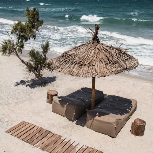 Two cushioned beach loungers under a thatched umbrella on a sandy beach with a small tree nearby and a wooden walkway leading to the loungers. Waves are visible in the background with distant islands on the horizon.