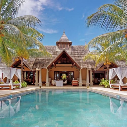 Eine luxuriöse tropische Villa mit einem großen Swimmingpool, Palmen und bequemen Sitzgelegenheiten im Freien unter überdachten Betten auf beiden Seiten des Pools.