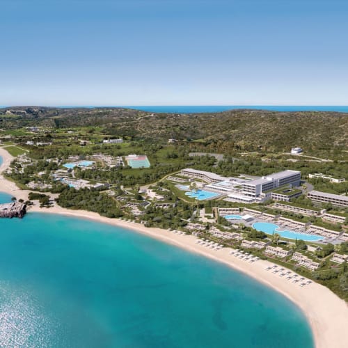 Panoramic view of a coastal resort with sandy beaches, swimming pools, and lush greenery, overlooking the blue sea.