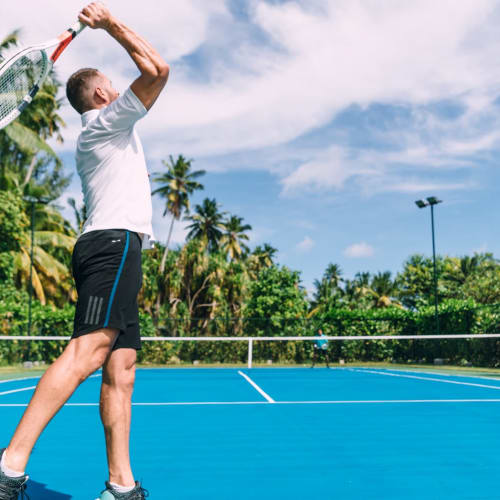 Ein Mann in Sportkleidung, der sich darauf vorbereitet, einen Tennisball auf einem blauen Tennisplatz zu schlagen, umgeben von üppiger tropischer Vegetation und Palmen unter teils bewölktem Himmel.