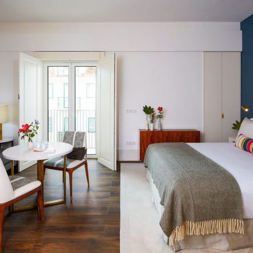 Innenraum einer modernen, hellen Hotelzimmer- oder Apartment-Wohnung mit Doppelbett, Sitzecke mit Sessel und Couch, Esstisch mit Stühlen, Balkon mit offener Tür, und dunkle Holzböden.